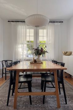 a dining room table with black chairs and a vase on top of it in front of two windows