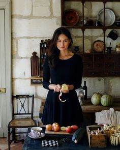 a woman in a black dress is holding an apple and standing next to some fruit on a table