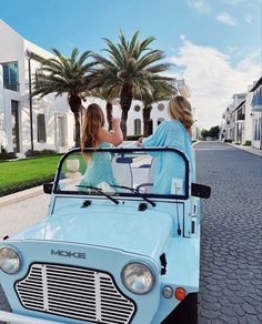 two women sitting in the back of an old blue car on a street lined with palm trees