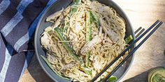 a bowl of noodles with chicken and asparagus on a wooden table next to chopsticks