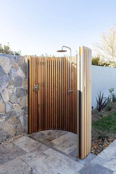 an outdoor shower with wooden slats on the side and stone wall behind it, in front of a white fence