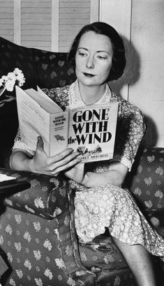 a woman sitting on a couch reading a book with flowers in the corner next to her