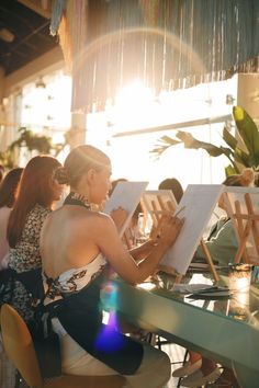 a woman sitting at a table with an easel and painting on the canvass