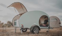 a man sitting in the back of a tear shaped camper trailer on dry grass