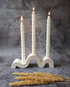 two white candles sitting next to each other on top of a gray surface with some dry grass