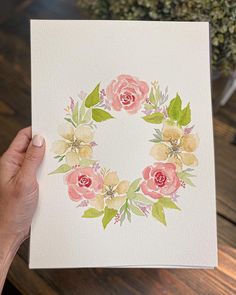 a hand holding up a card with watercolor flowers and leaves in the shape of a wreath