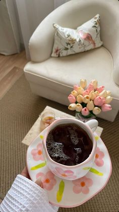 a person holding a coffee cup and saucer in front of a white chair with flowers on it