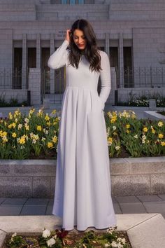 a woman standing in front of a building wearing a long white dress and holding her hair