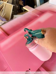 a hand is holding a spray bottle with pink paint on it and there are other items in the background