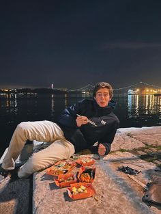 a man sitting on the edge of a pier eating food at night with lights in the background