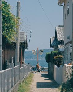 a person riding a bike down a street next to houses and water with mountains in the background