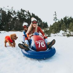 two people are riding on a tube in the snow with a dog following behind them