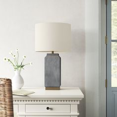 a white table with a lamp and vase on it next to a wicker basket