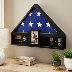 an american flag is hanging on the wall next to a shelf with two pictures and a potted plant