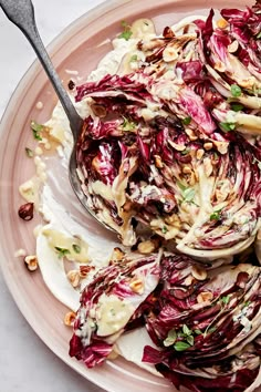 a pink plate topped with red cabbage and shaved nuts next to a spoon filled with dressing