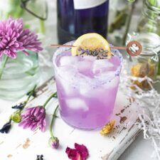 a purple drink sitting on top of a wooden table next to flowers and bottles filled with liquid