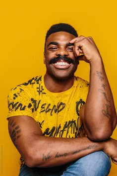 a man with tattoos and a moustache sitting in front of a yellow wall