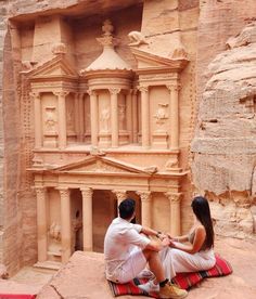 a man and woman sitting on the ground in front of a building carved out of rock