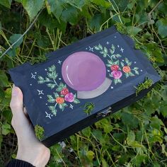 a hand holding a painted box with a purple plate on it and flowers around the lid