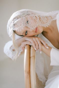 a woman wearing a white dress and veil leaning on a wooden chair with her hand on the back of it