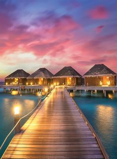 a pier leading to over water huts with lights on them at dusk in the ocean