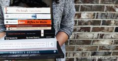 a woman holding a stack of books in front of a brick wall with her hands on top of them