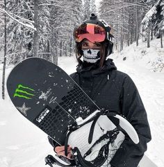 a snowboarder is holding his board while standing in the snow with trees behind him