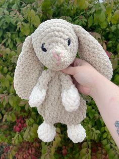 a hand holding a small crocheted bunny in front of some bushes and plants