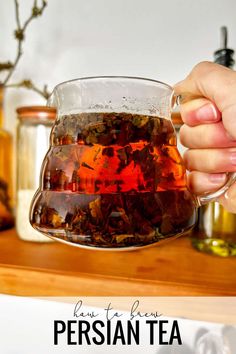 a person holding a glass cup filled with tea on top of a wooden shelf next to jars