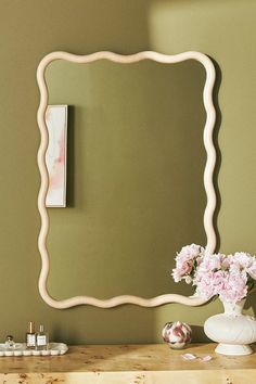 a white vase with pink flowers sitting on top of a wooden table next to a mirror