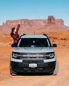 a woman standing on the hood of a silver truck in front of mountains and desert