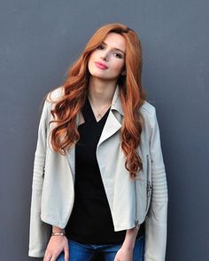 a woman with long red hair standing in front of a gray wall wearing a white jacket and jeans