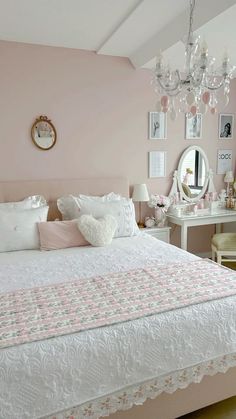 a white bed sitting under a chandelier next to a mirror and table in a room