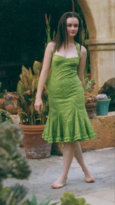 a woman in a green dress is standing near some potted plants and looking at the camera