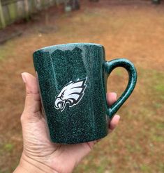 a hand holding a green coffee mug with the philadelphia eagles on it in front of a yard