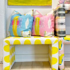 three colorful pillows sit on a white bench in front of a wall with yellow and pink accents