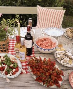 an outdoor table with plates and bowls of food on it, bottles of wine in the background