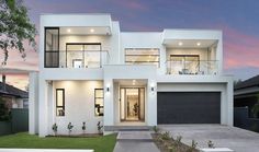 a modern house with white walls and grass in front of it at dusk, as seen from the driveway