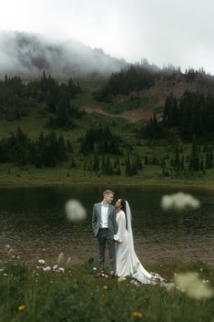 a bride and groom standing in front of a lake surrounded by wildflowers on a cloudy day