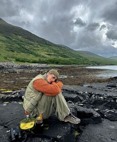 a man sitting on top of a rocky beach next to a body of water with mountains in the background