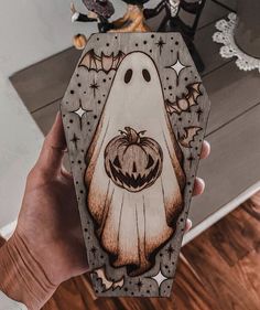 a hand holding up a wooden box with a ghost on it's face and pumpkins in the background