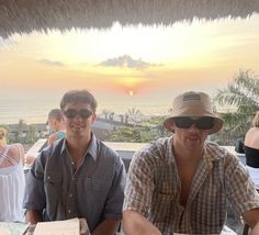 two men sitting at a table with pizza in front of them and the sun setting behind them