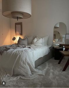 a bed with white sheets and pillows in a bedroom next to a table, mirror and lamp