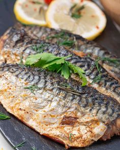 two grilled fish on a black plate with lemons and herbs next to it