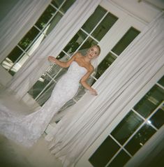 a woman standing in front of a window wearing a wedding dress and holding her hand out