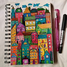 an open notebook with colorful cityscape drawn on it next to two marker pens