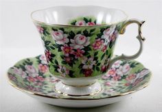 a tea cup and saucer decorated with pink flowers on a green floral pattern, gold trimming around the edge