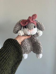 a hand holding a gray and white crocheted stuffed animal with a pink bow