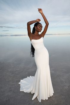 a woman in a white dress is standing on the beach with her arms up and hands behind her head