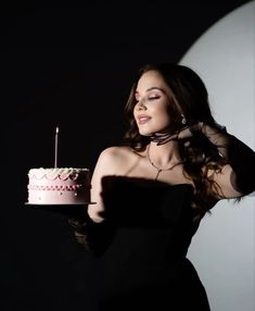 a woman in a black dress holding a cake with a candle on it and looking at the camera